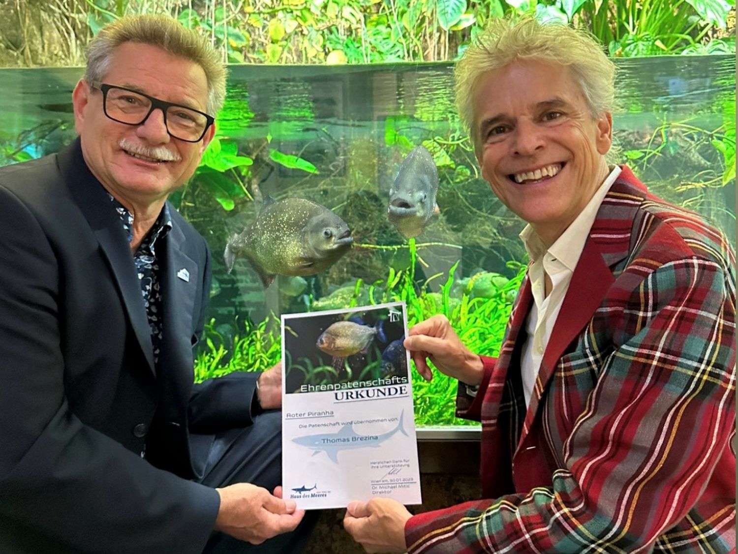 Thomas Brezina und ehemaliger Direktor Michael Mitic halten die Urkunde für die Piranha-Patenschaft in die Kamera